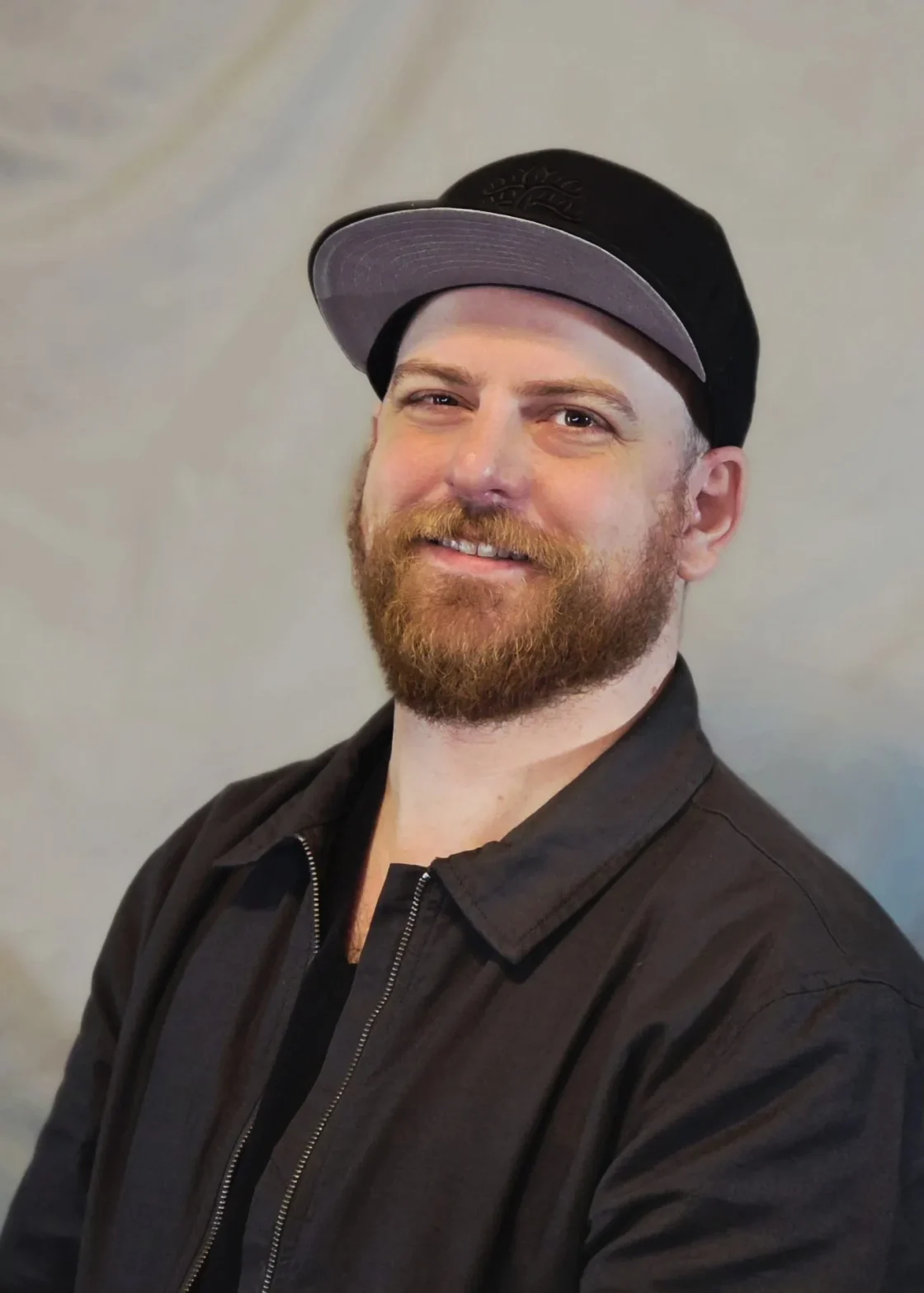 Professional headshot of bearded man in black jacket and baseball cap for food service industry business networking.